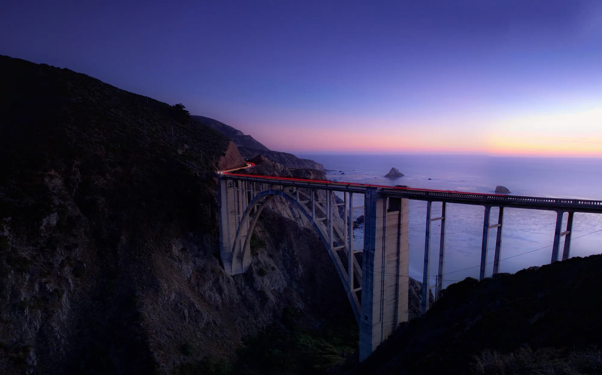 Bixby Creek Bridge spanning a coastal canyon at dusk, car light trails crossing toward the darkening Pacific